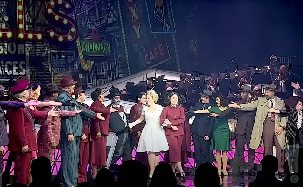 A cast of performers in vintage attire on stage, with a band in the background and a cityscape backdrop.