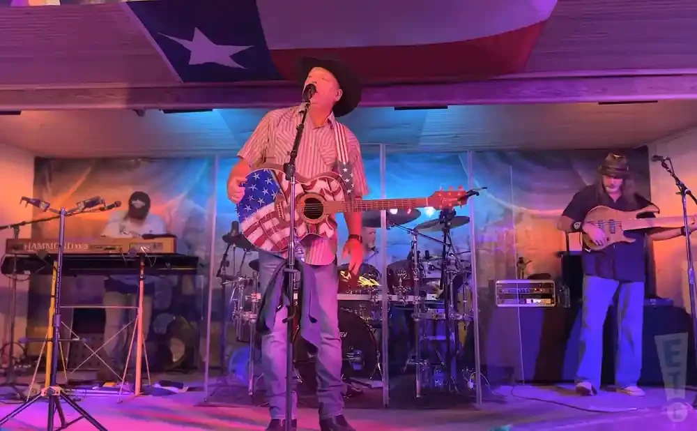 A man in a cowboy hat plays an American flag-decorated guitar while a band performs on stage.