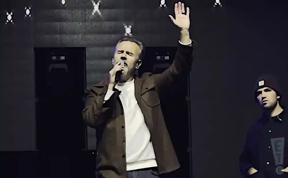 A man with a microphone and raised hand sings on stage, with another person in a beanie standing to his right.
