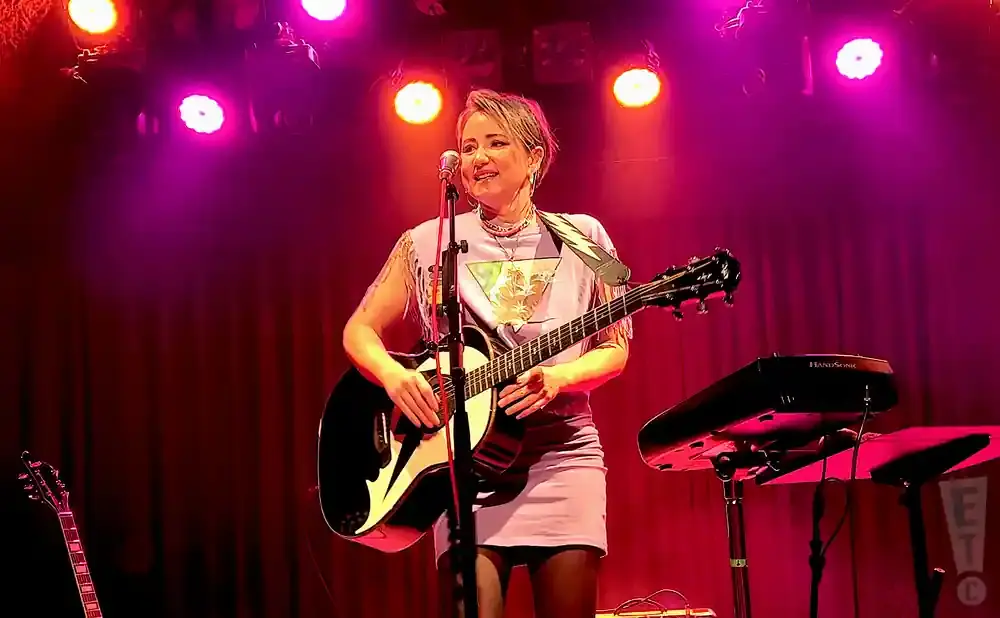 Woman plays acoustic guitar on stage with pink and purple lights.