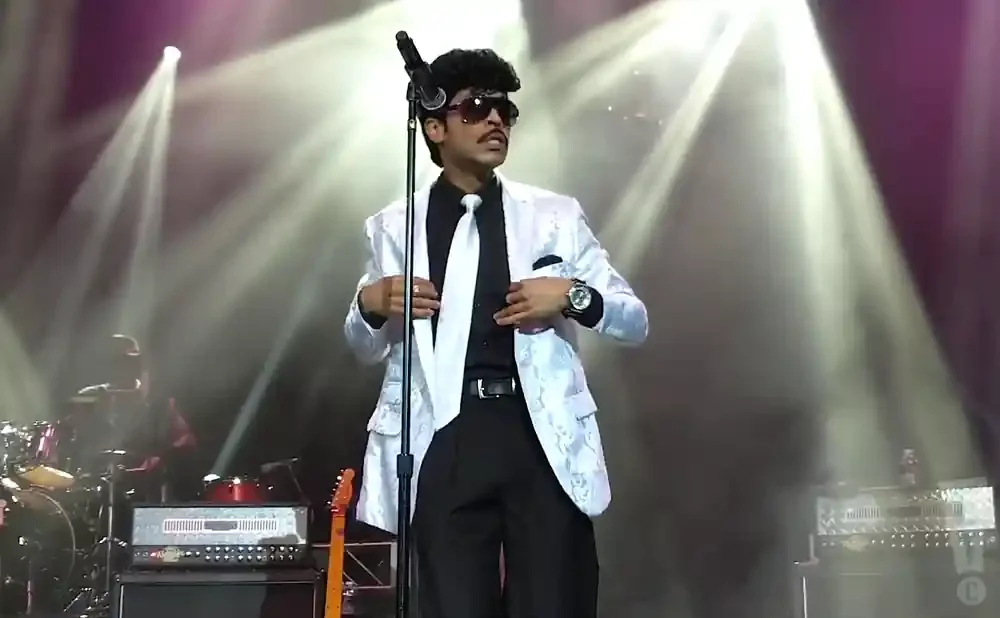 Performer in a white suit and tie sings into a microphone, with stage lights in the background.