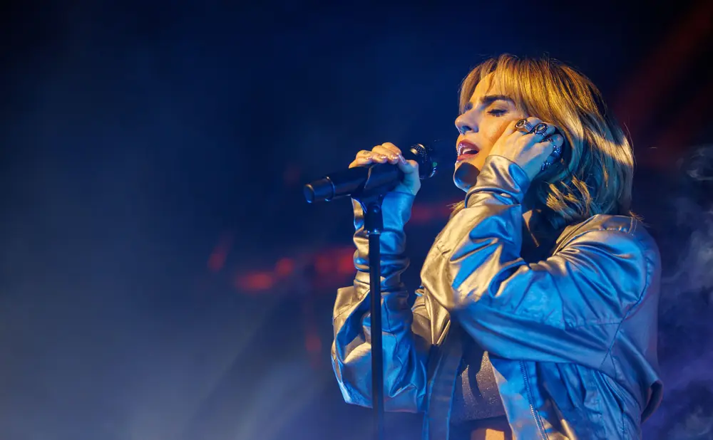 A woman sings into a microphone on stage, her eyes closed and hand to her cheek, illuminated by stage lights.