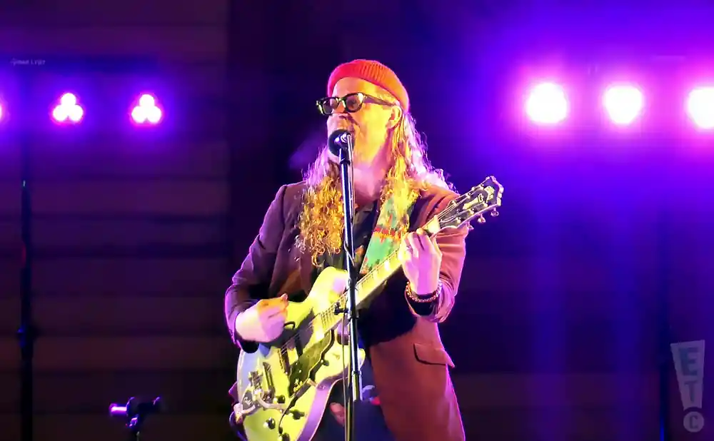 Musician with curly blonde hair, wearing a red beanie and glasses, plays a light green guitar under purple stage lights.