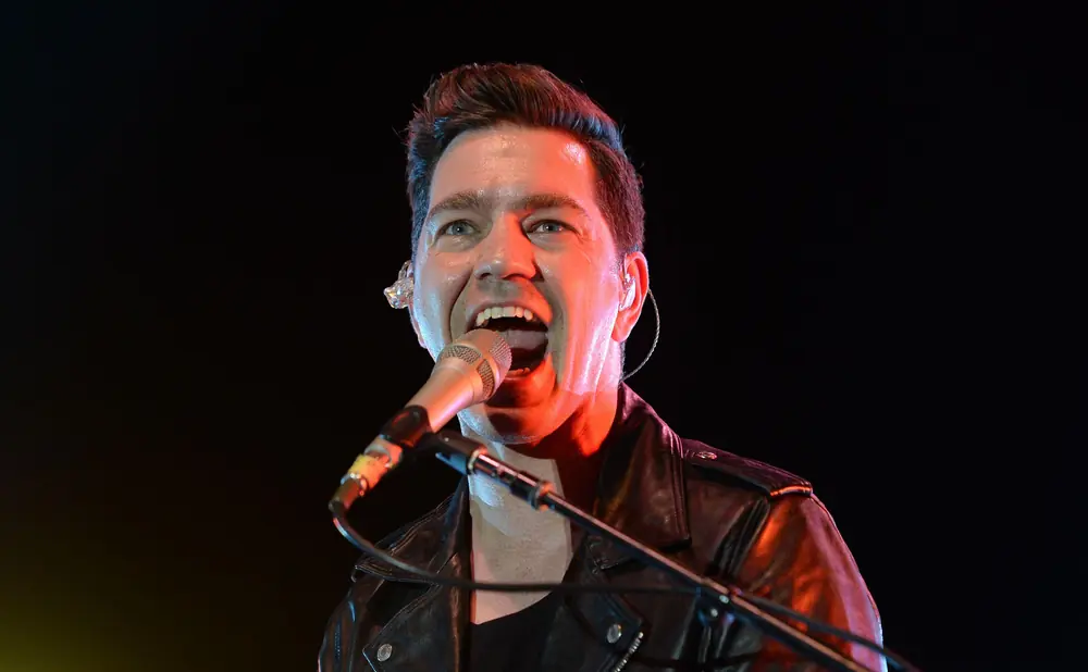 A man in a leather jacket sings into a microphone on a dark stage, lit by red and yellow lights.