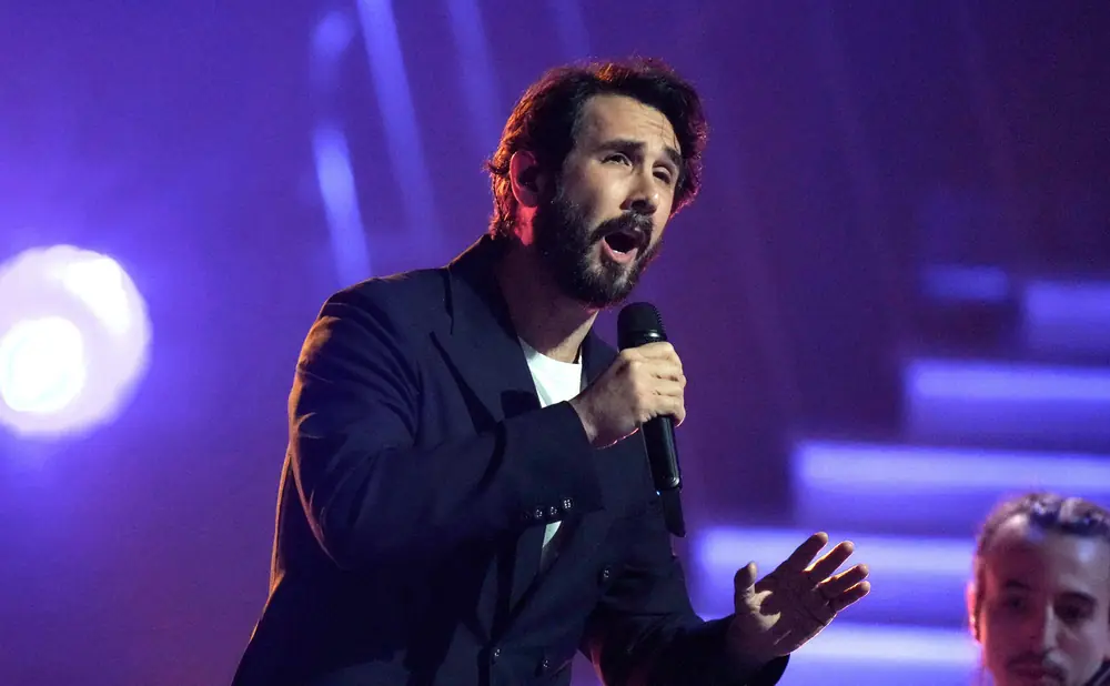 Singer on stage with a sparkly jacket, singing into a microphone, with a large crowd and confetti.