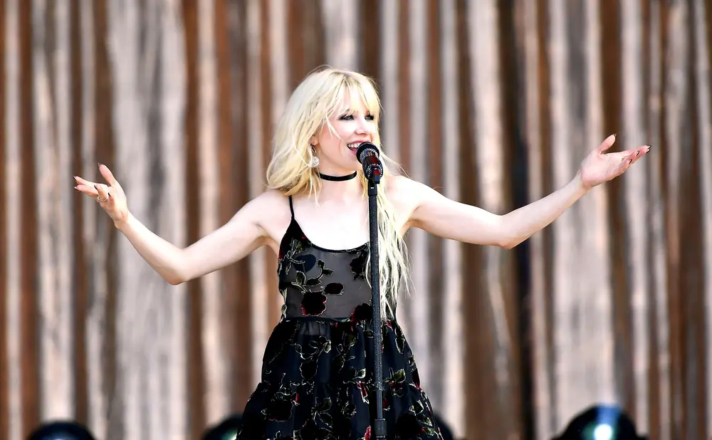 A blonde woman in a floral dress sings into a microphone on stage.