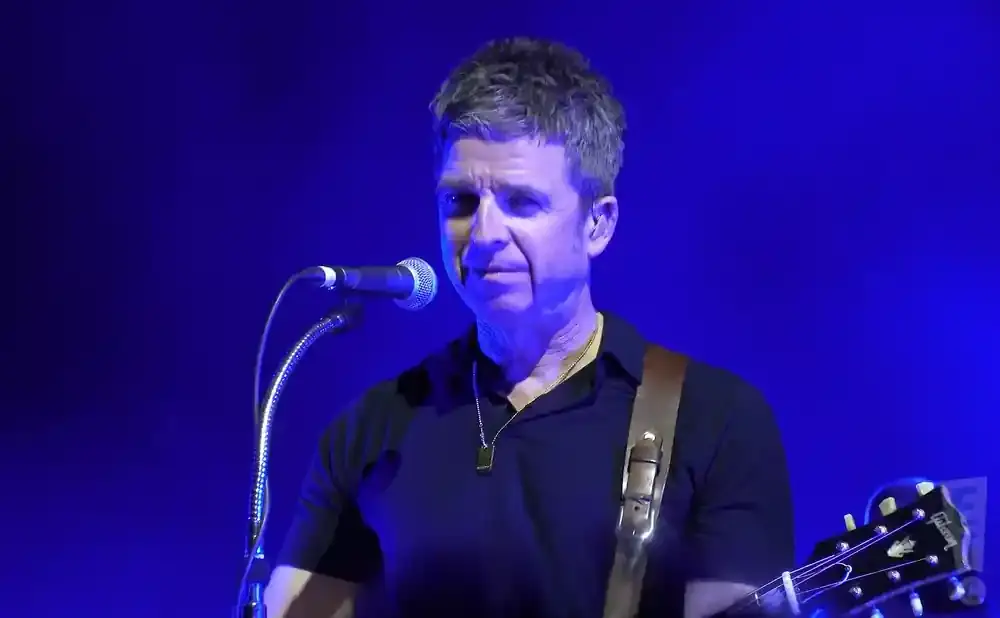 Noel Gallagher on stage playing guitar, wearing a black polo shirt and a dog tag necklace, with a blue background.