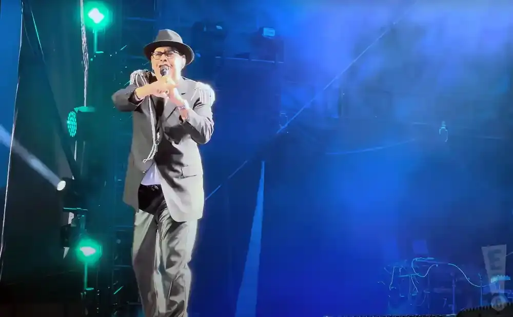 A man in a black hat and suit performs on a stage with blue and green lights.