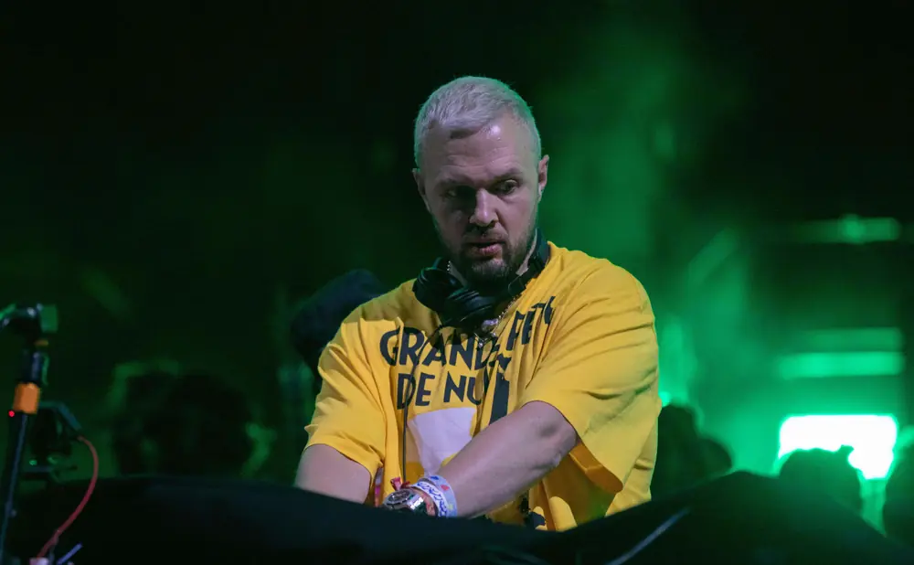A DJ with blond hair and a yellow shirt plays music on a turntable under green lights.