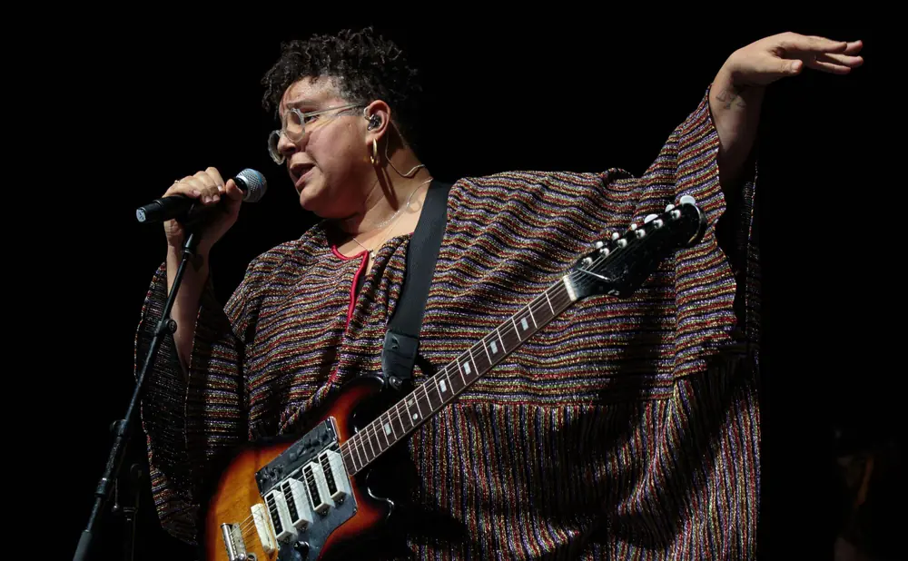 A singer performs on stage in a stadium with a large crowd holding up lights and fireworks in the background.