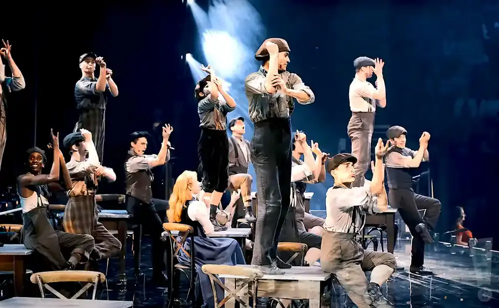 A diverse group of performers in historical costumes dance and sing on a stage with bright lighting.
