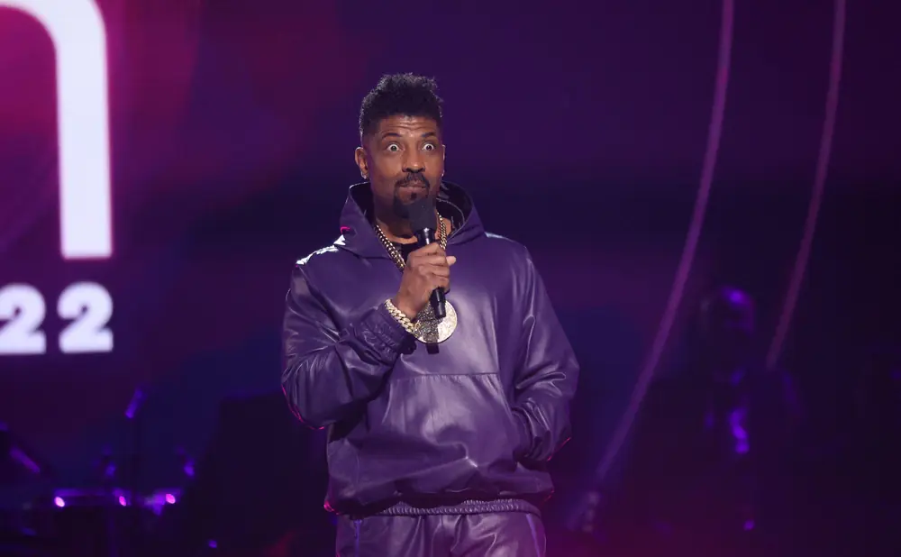 Comedian Deon Cole on stage in a purple leather tracksuit and gold chains.