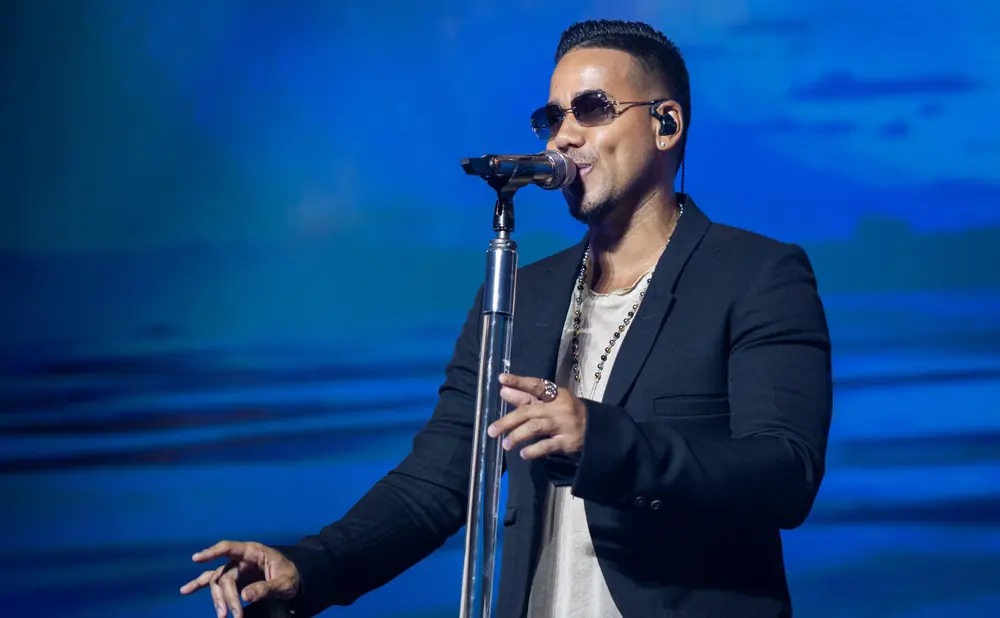 A man in a white shirt and pants sings into a microphone on a dimly lit stage with purple lights.