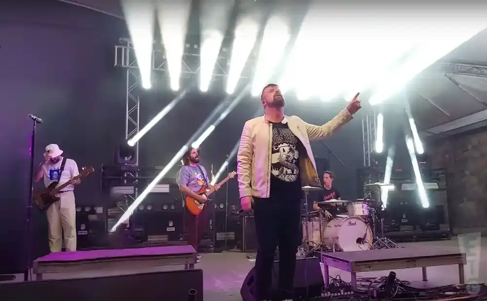 A band performs on stage with bright lights. The lead singer points up.
