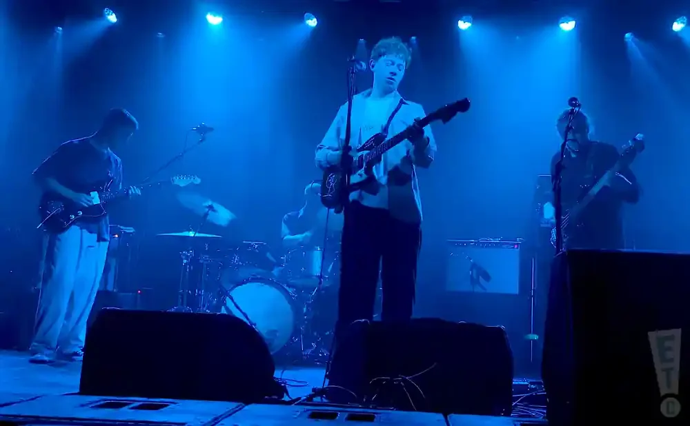 A band performs on a dimly lit stage under blue lights, with three guitarists and a drummer.