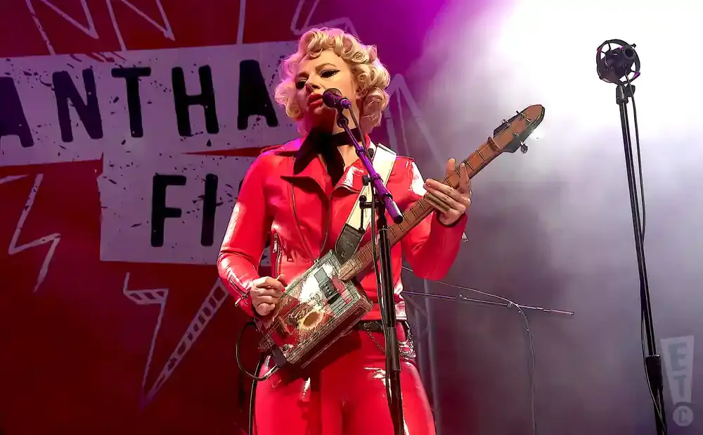Woman in a red leather outfit plays a cigar box guitar on stage, with a microphone in front of her.