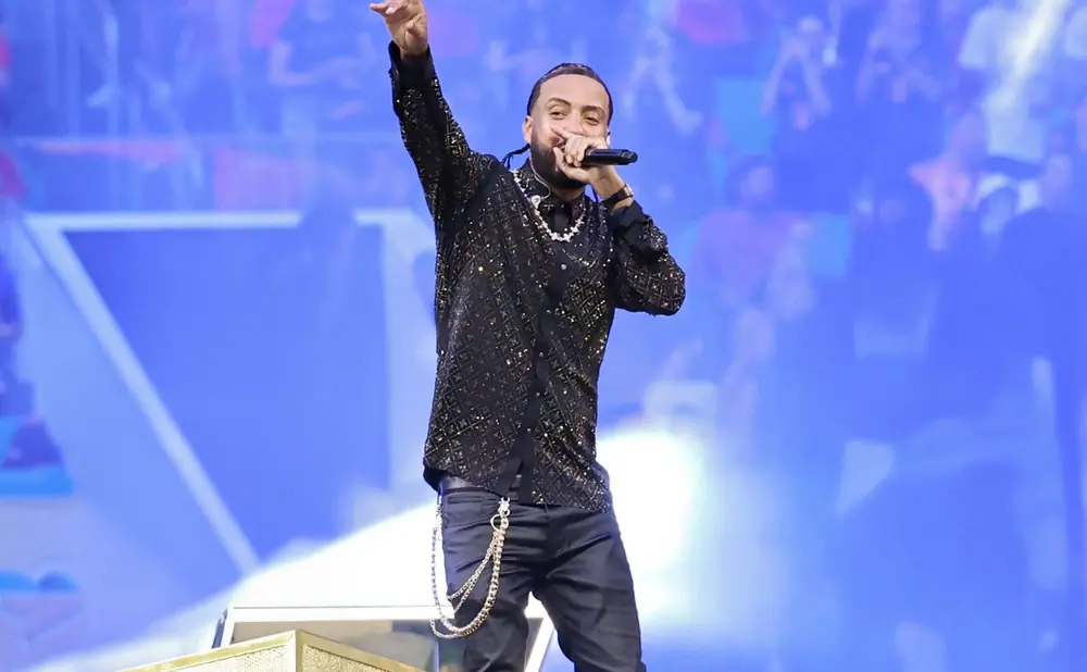 A rapper with braided hair, wearing a black sequined shirt and black pants with a gold chain, performs on stage with a microphone.
