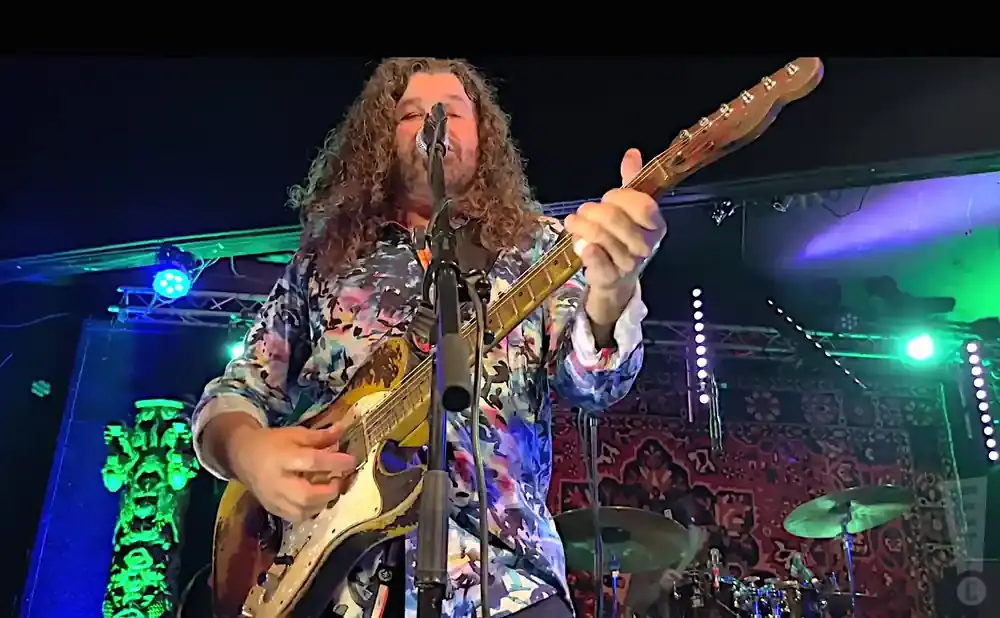 A man with long curly hair plays a guitar on stage, wearing a colorful patterned shirt.