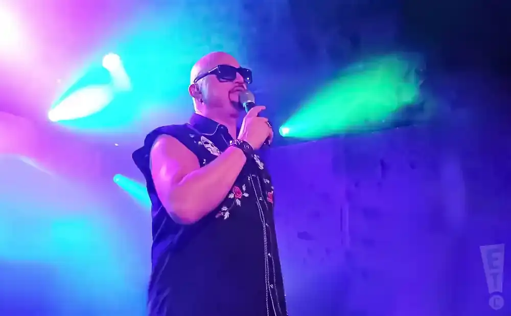 Bald man in sunglasses and black embroidered shirt sings into a microphone on a stage with colorful lights.