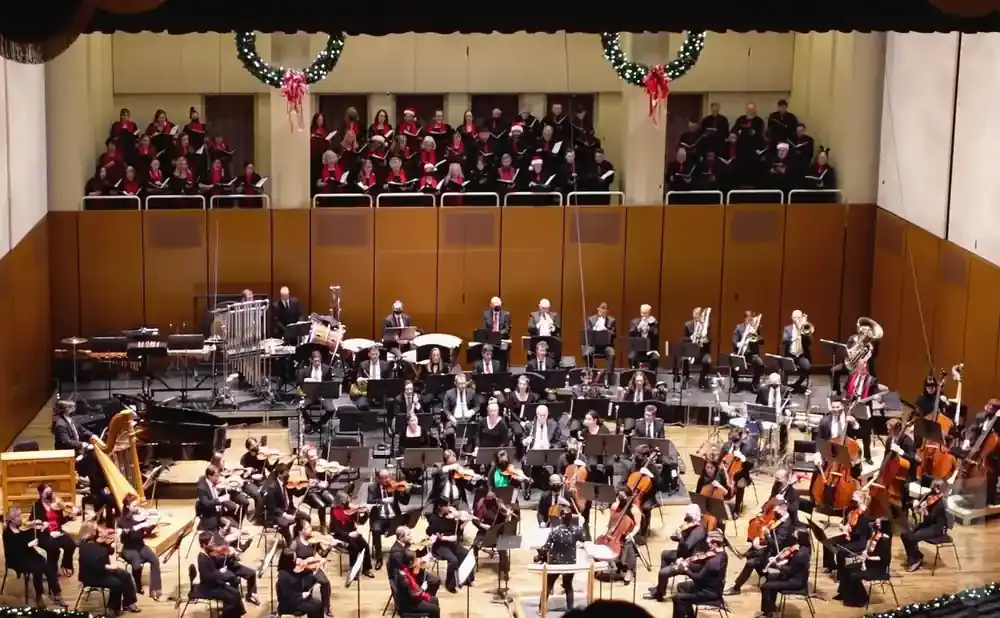 An orchestra performs on stage with a choir in the background, decorated for the holidays.