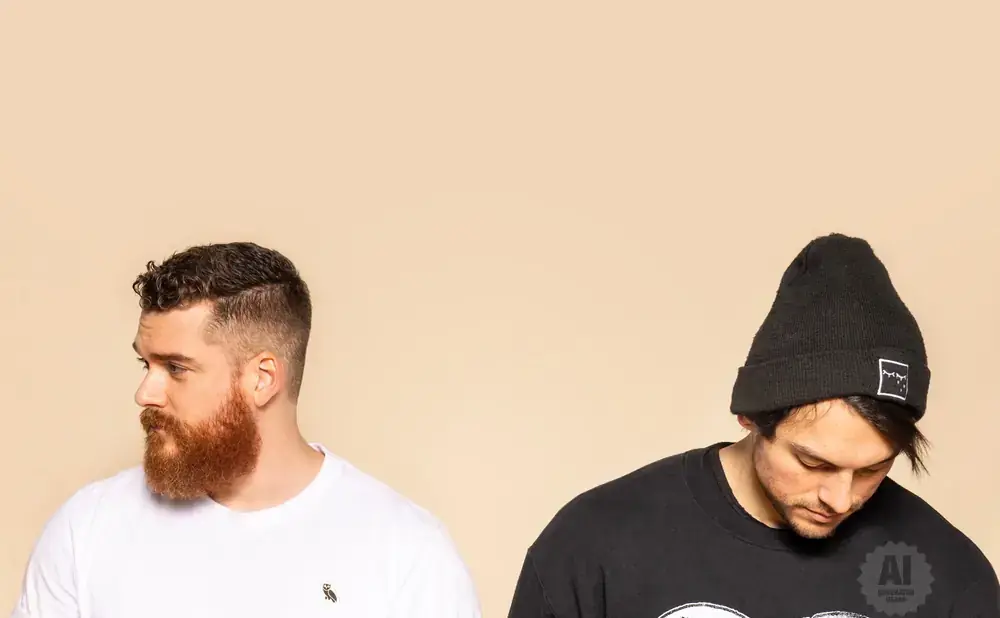 Two men against a plain background: one with a beard in a white t-shirt, the other with a beanie in a black sweatshirt.
