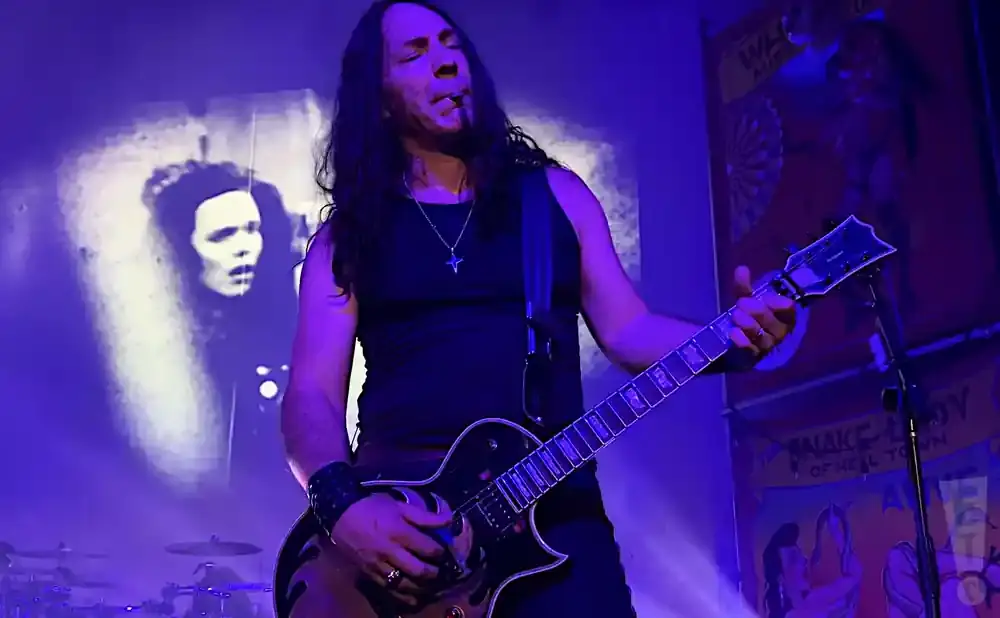 A man with long dark hair plays a black electric guitar, smoking a cigarette, on a dimly lit stage.