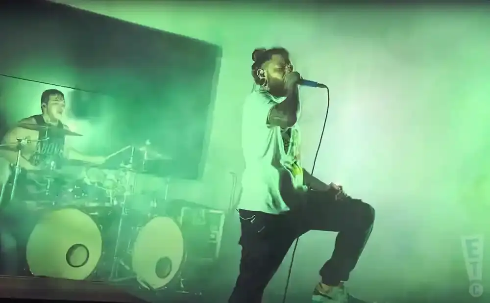 A singer on stage holds a microphone, while a drummer plays behind him, bathed in green stage lights.