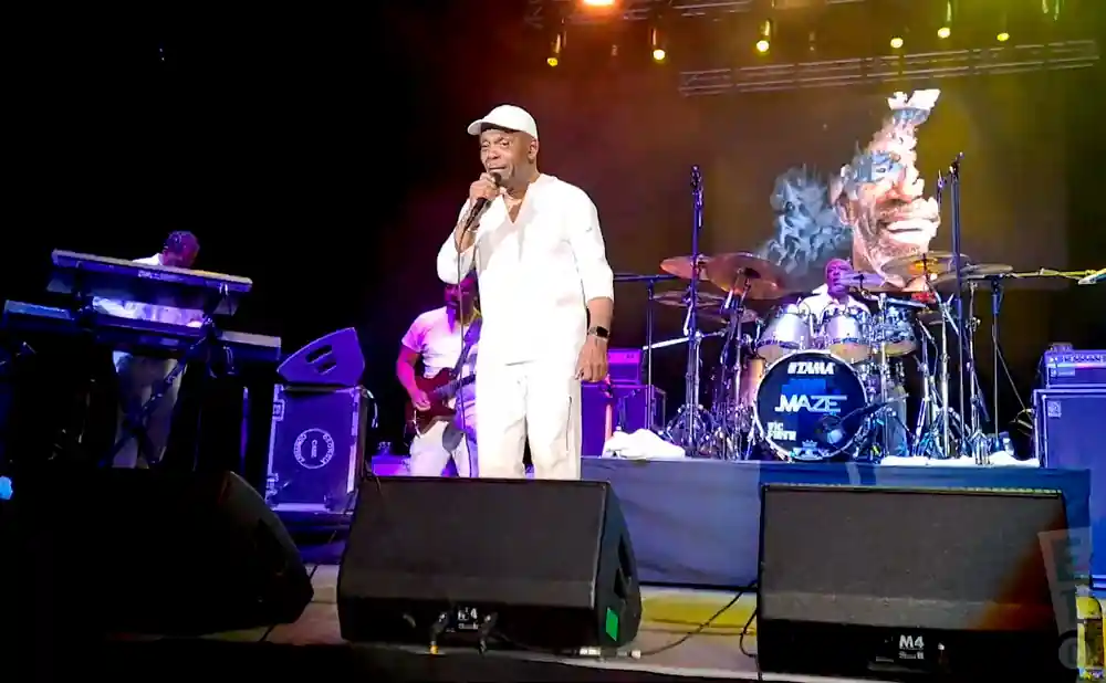 Musician in white on stage singing into a microphone with a band playing behind him.