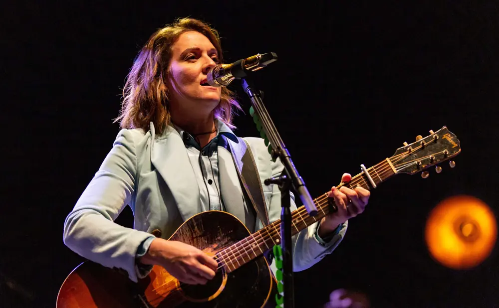 A woman sings and plays an acoustic guitar on stage, lit by stage lights against a dark background.