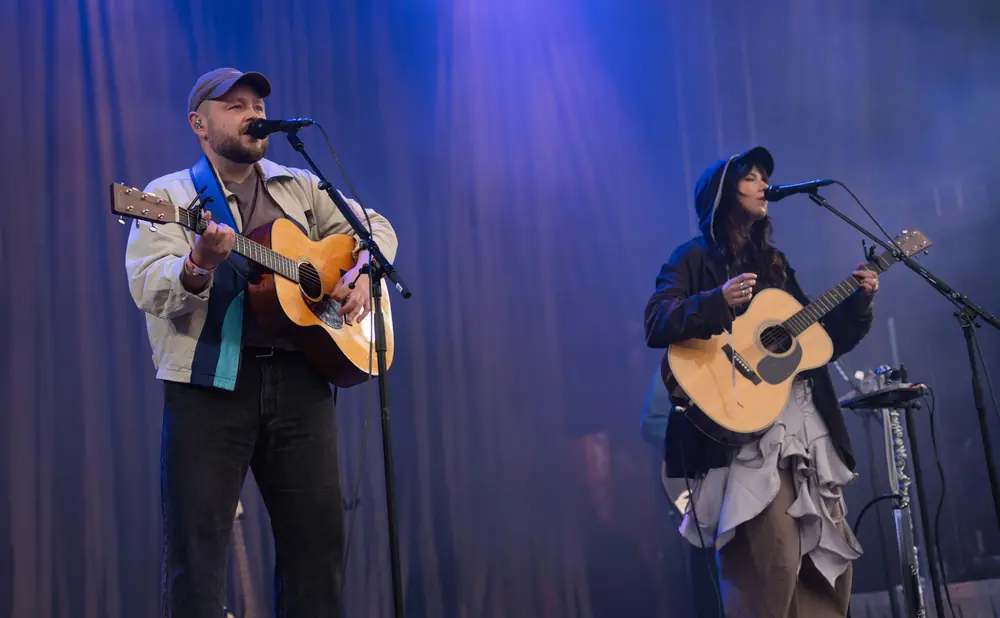 Musicians on stage with blue lighting: a man plays guitar and sings into a microphone, a woman plays guitar.