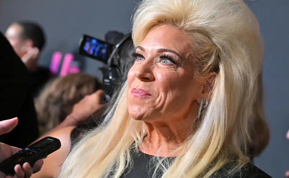 Theresa Caputo with blonde hair, wearing a black dress and sparkling earrings, speaks into a microphone.