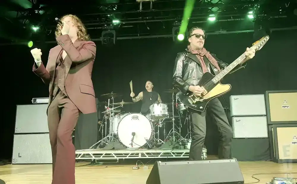 Musicians perform on stage, with a singer in a suit on the left, a guitarist in a leather jacket on the right, and a drummer in the background.