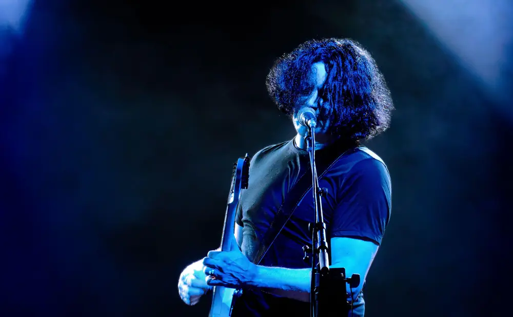 A musician with dark, curly hair plays a guitar under blue stage lights.