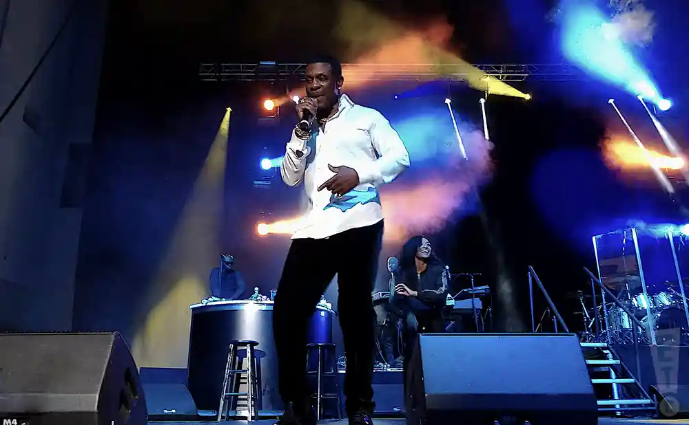 A man in a white shirt and black pants sings into a microphone on a stage with colorful lights.