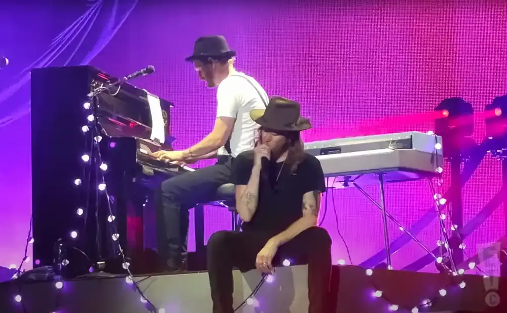 Two men on a stage, one playing piano, the other singing into a microphone, both wearing hats.