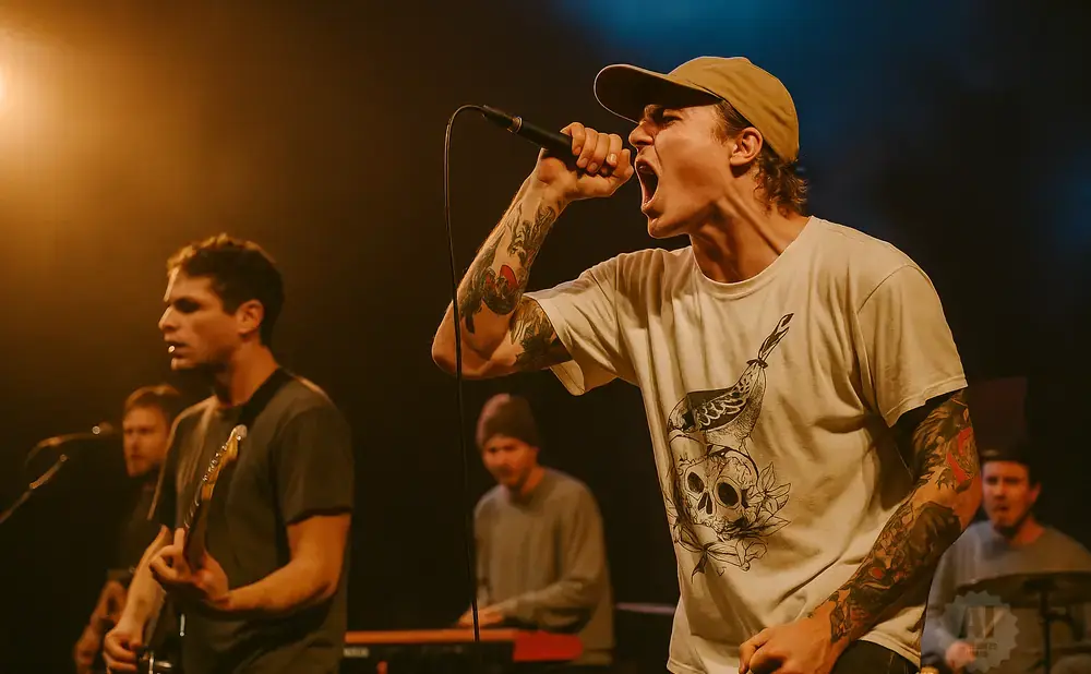 A male singer with a cap and tattoos sings into a microphone on stage, with bandmates behind him.