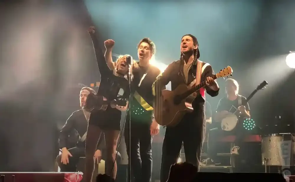 Musicians sing and play instruments on a brightly lit stage, with a female singer in the foreground raising her fist.