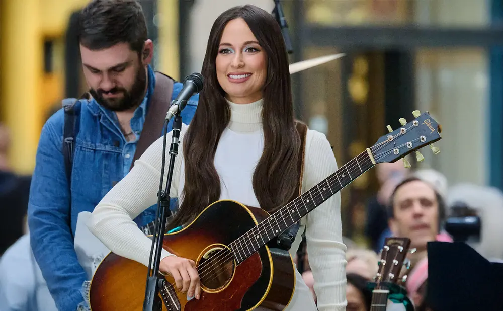 Singer Kacey Musgraves plays an acoustic guitar on stage, wearing a white turtleneck and brown vest.