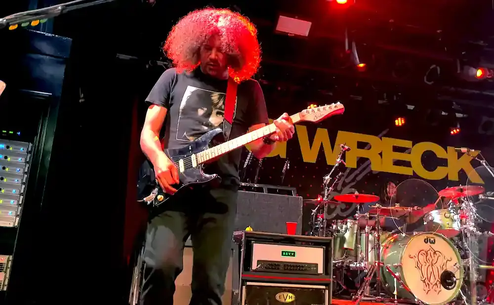 A guitarist with a large afro plays a black electric guitar on stage, with a 