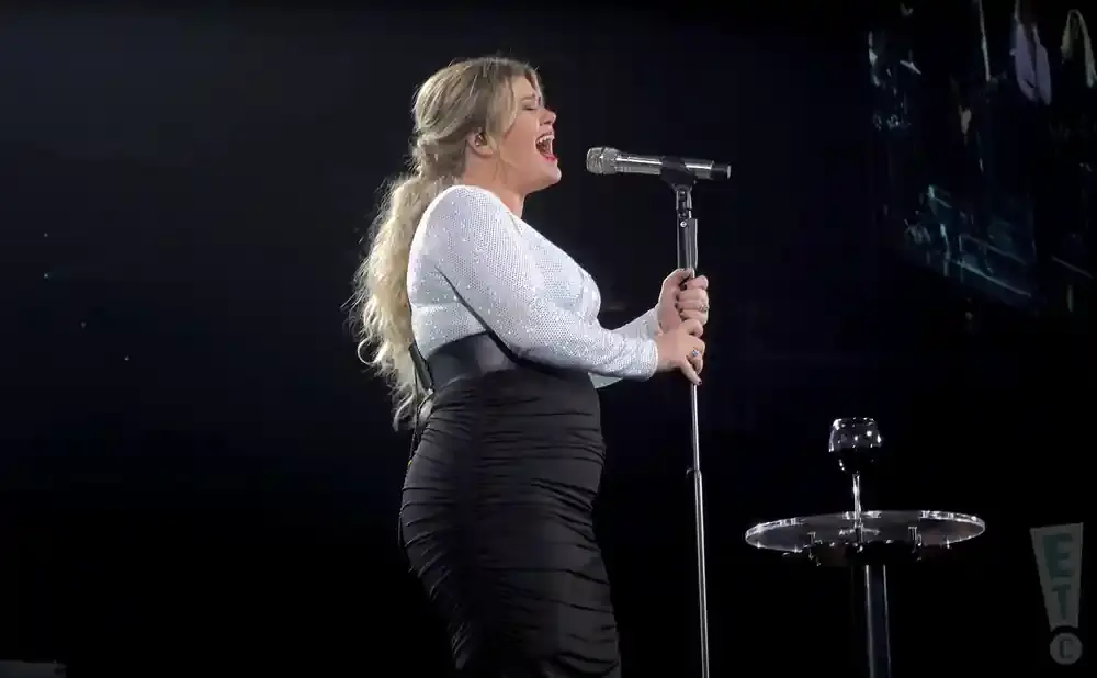 Singer Kelly Clarkson performs on stage wearing a sparkly white top and a ruched black skirt.