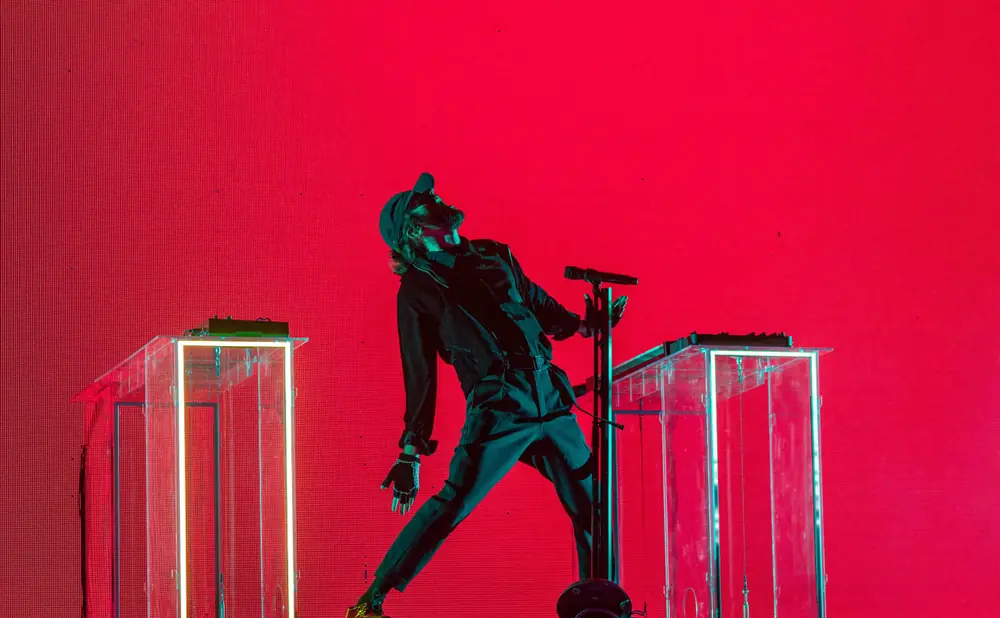 A singer in a black outfit and cap performs on a stage in front of a bright red LED screen.