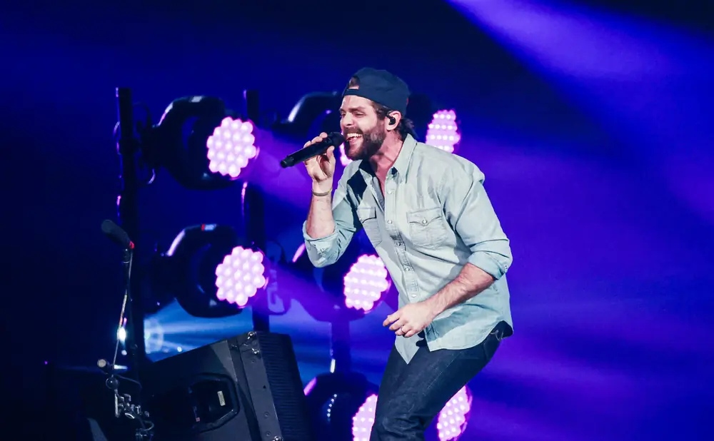 Man in a baseball cap and denim shirt sings into a microphone on stage with purple lights.