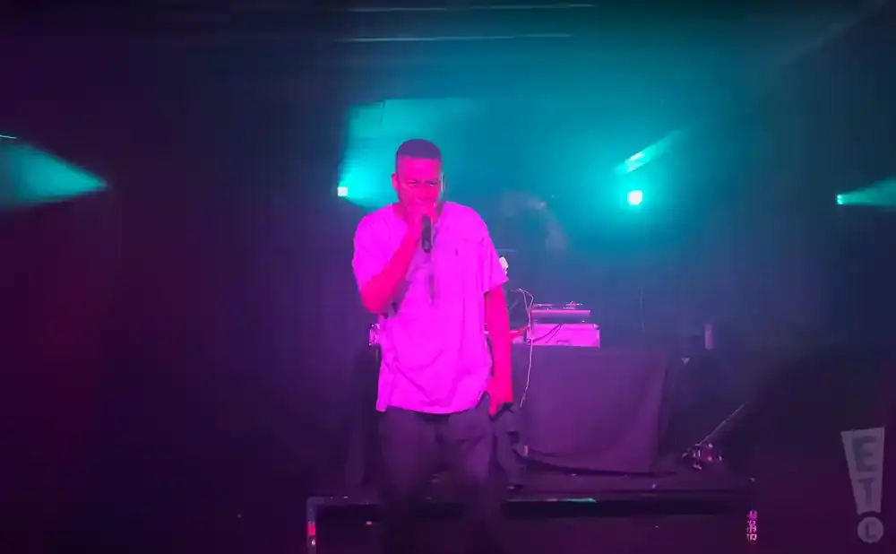 Rapper performing on a dimly lit stage with purple and teal spotlights.