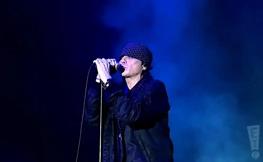 A man with a bandana on his head sings into a microphone on a dark, smoky stage lit by blue lights.