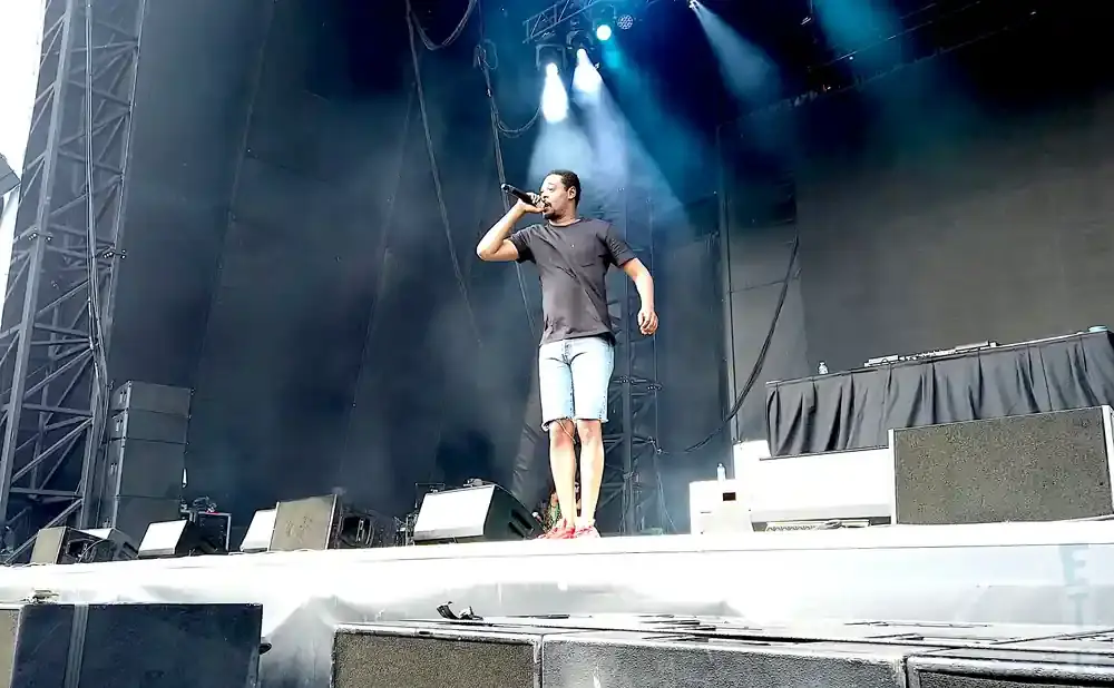 A man in a black t-shirt and denim shorts performs on stage with a microphone.