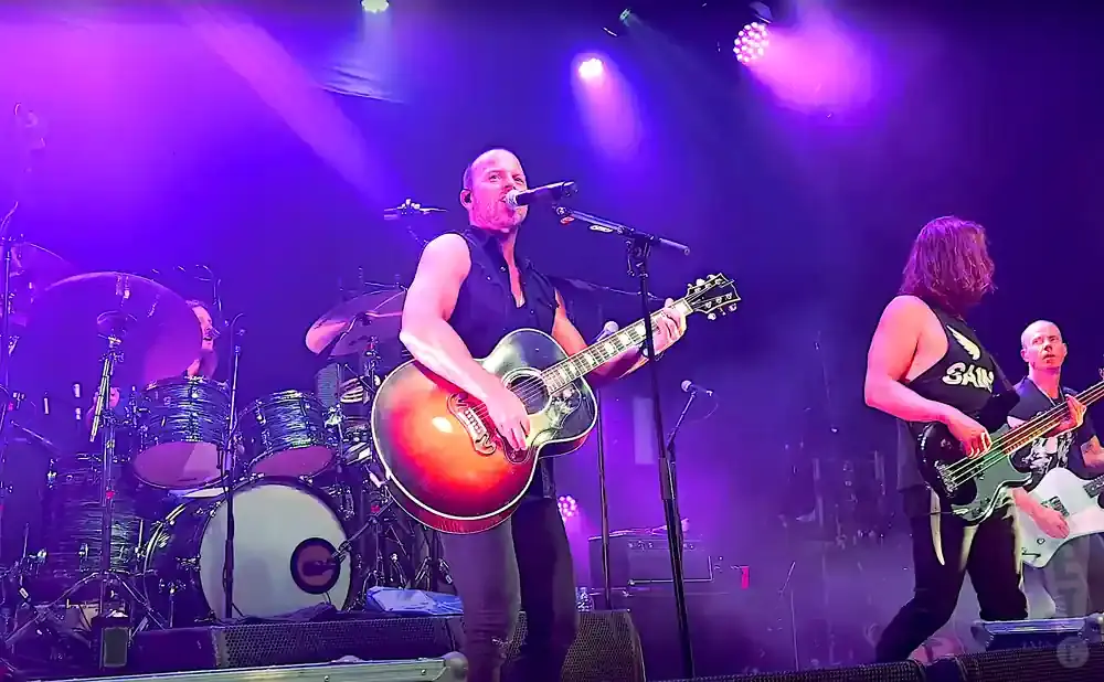 A bald man with a sleeveless shirt plays an acoustic guitar and sings into a microphone on a stage with purple lighting.