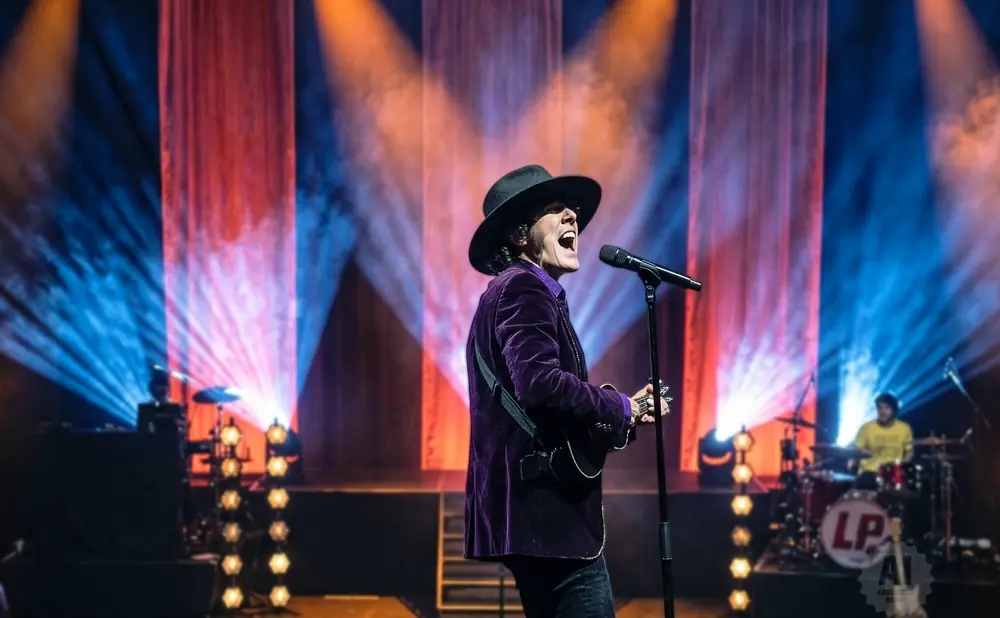 Singer LP performs on stage wearing a black hat and purple velvet jacket, with a microphone and guitar.
