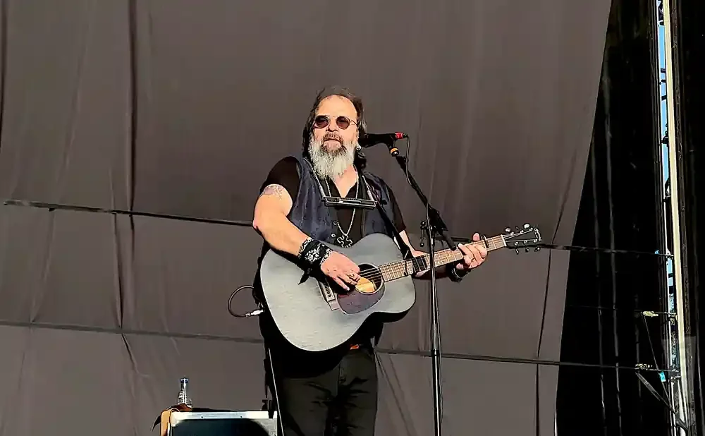 A man with a beard and sunglasses plays an acoustic guitar on stage.