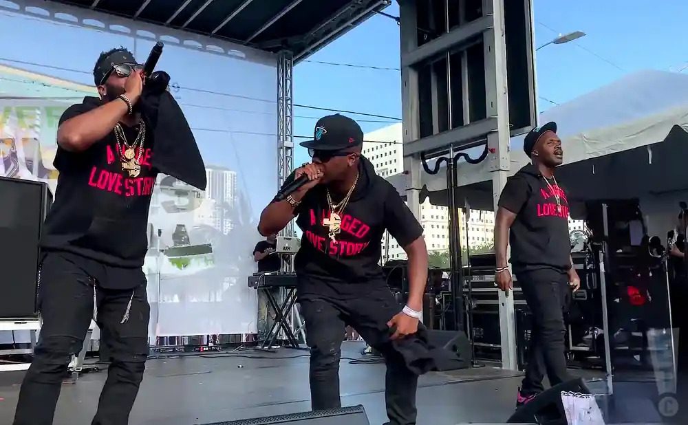 Three men in black hoodies and pants perform on a stage.