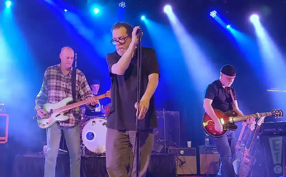 A band performs on stage with blue lights, featuring a singer, bassist, and guitarist.